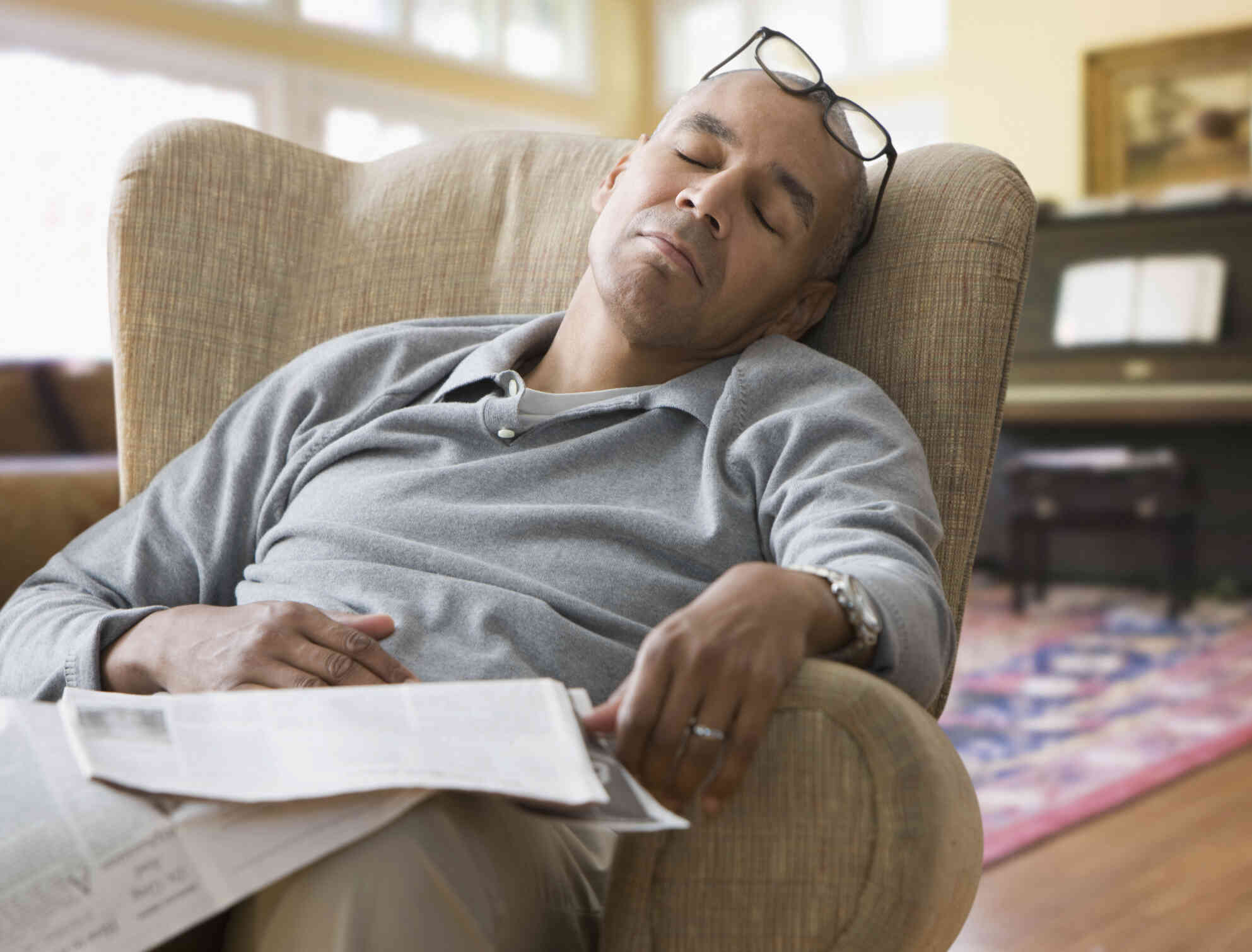 A middle aged man sleeps in an armchair in his home with his classes on his forehead and a mewspaper in his lap.